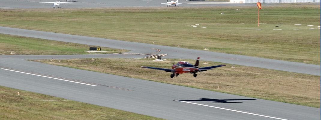 Piper Archer landing at small rural airfield with wind sock, other aircraft, and taxiway visible