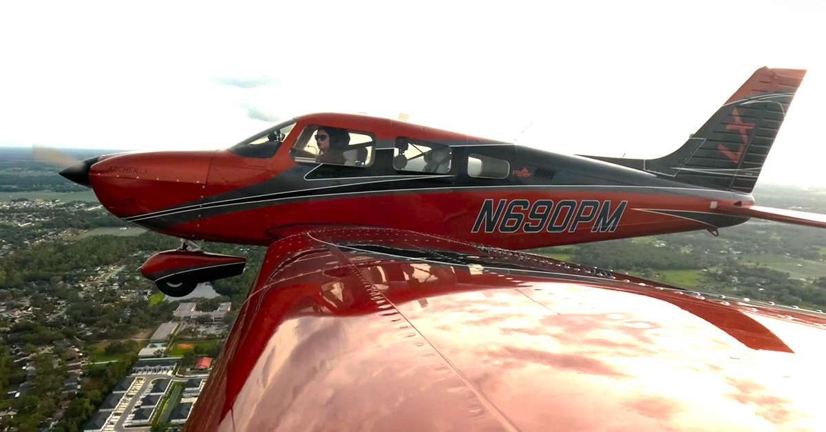 Female pilot seen flying in side-view photo of Piper Archer over landscape in summer