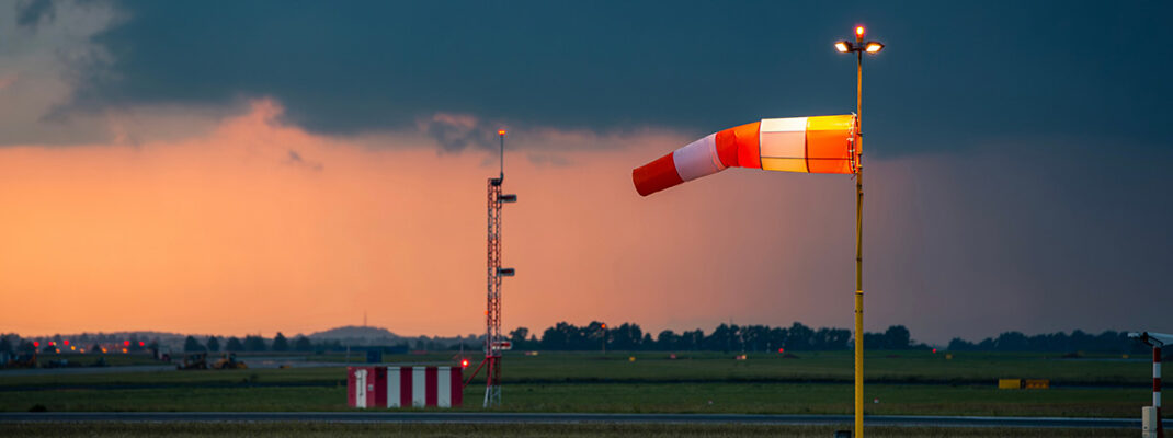 Red and white striped windsock fully extended by strong wind at airport airfield against oncoming dark storm clouds.