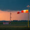 Red and white striped windsock fully extended by strong wind at airport airfield against oncoming dark storm clouds.