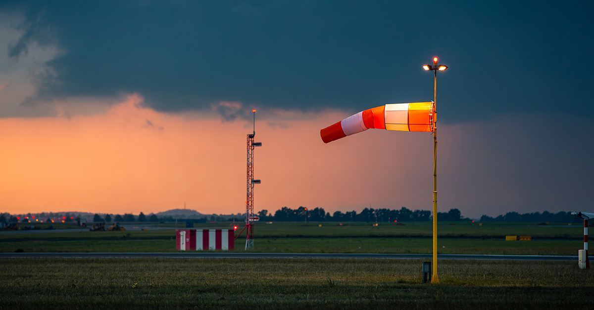 Red and white striped windsock fully extended by strong wind at airport airfield against oncoming dark storm clouds.