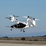 An experimental eVTOL aircraft above runway during take-off with mountains behind.